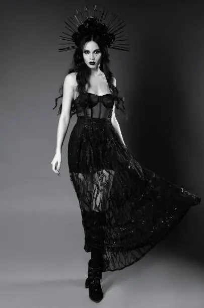 Photo of Halloween theme: attractive young witch in black dress and headgear with roses and spikes. Dark beauty: portrait of seductive cute goth girl. Black and white