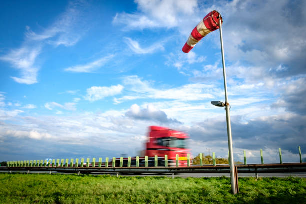 Truck driving past a wind sock near a highway during a windy day with blue sky and clouds in the background. The truck is blurred because of the speed it is driving at.