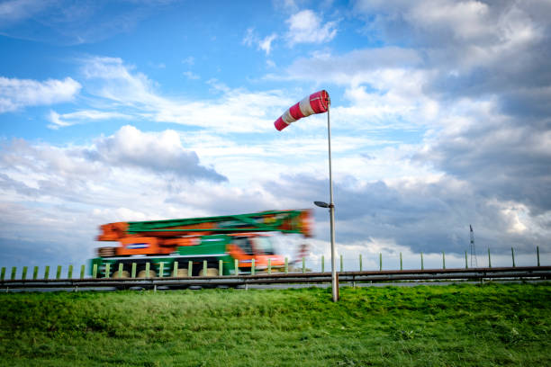Truck driving past a wind sock near a highway during a windy day with blue sky and clouds in the background. The truck is blurred because of the speed it is driving at.