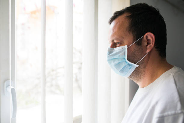 istock Lonely man in medical mask looking through the window. Isolation at home for self quarantine 1279784218