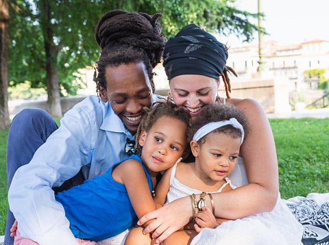 family united in a tender and loving embrace, African father with Caucasian mother hold and cuddle their children in a public park, portrait of rastafarian family, diversity and multi-ethnic concept