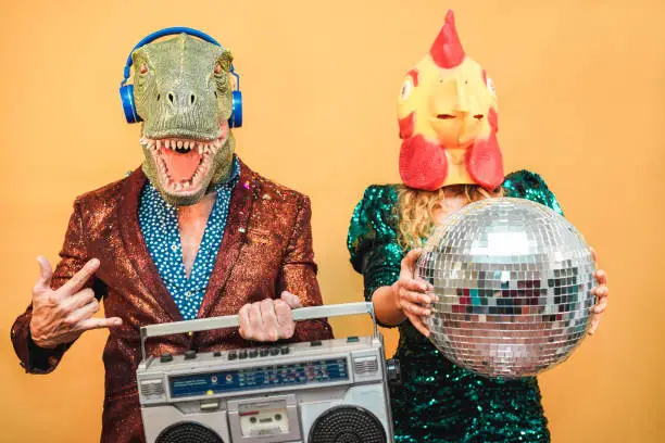 Photo of Crazy stylish people listening music with vintage boombox stereo - Fashion couple wearing t-rex and chicken mask at party fest event -  Absurd, holidays and funny trend concept - Focus on man face