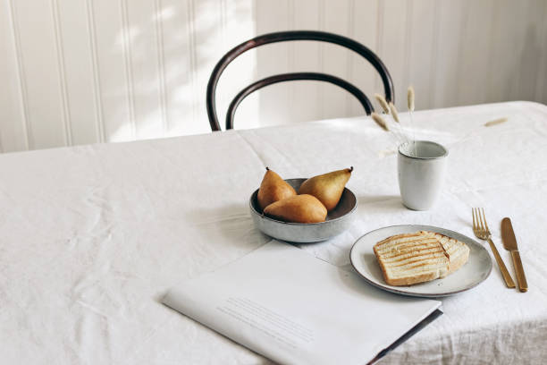 Morning breakfast with pears fruit composition. Bread toast, folded newspapers and dry grass in mug on white linen table cloth, minimal Scandinavian interior, dinning room in sunlight. Nordic kitchen.