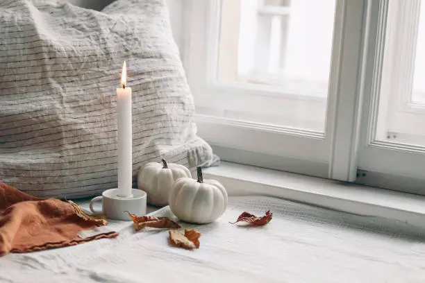 Autumn still life. Burning candle in white ceramic candleholder. Dry beech leaves and linen pillow near window. Moody composition with white pumpkin, Scandinavian interior.Thanksgiving, fall concept