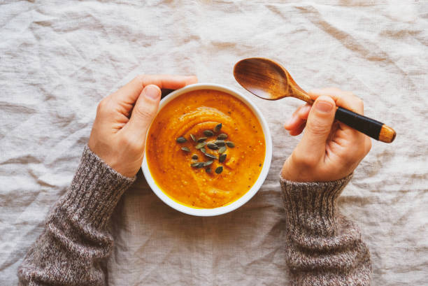 Male hands holding a bowl of hot pumpkin soup served with squash seeds on white tablecloth background, from above. Flat lay. Homemade autumn food. Popular Thanksgiving dish. Male hands holding a bowl of hot pumpkin soup served with squash seeds on white tablecloth background, from above. Flat lay. Homemade autumn food. Popular Thanksgiving dish. soup stock pictures, royalty-free photos & images