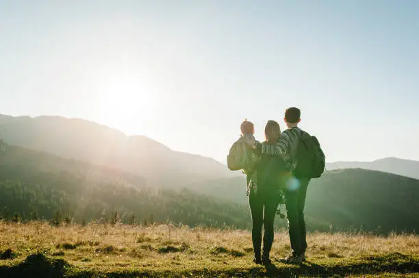 Photo of The daughter hug parents on nature. Mom, dad, and child walk in the grass. Family spending time together in mountain outside, on vacation. Family holiday trip concept. World Tourism Day. Back view.