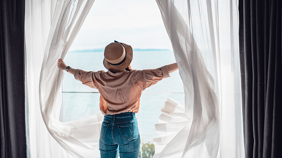 La mujer con vista trasera que abre cortinas blancas disfruta de la vista al mar, feliz estancia de viajero en un hotel de alta calidad. photo