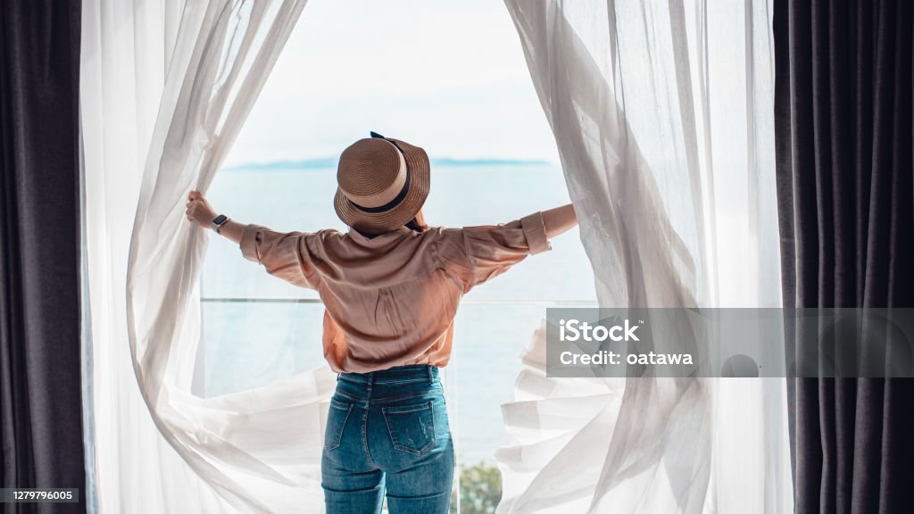 Rear back view woman opening white curtains enjoy sea view, Happy traveller stay in high quality hotel. Hotel Stock Photo