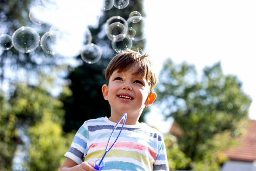 Three years boy in the park blowing soap bubbles and having fun. Brown hair kid in colorful shirt blows bubbles in the garden. Close up portrait of young toddler blowing soap bubbles in park. Copy-space