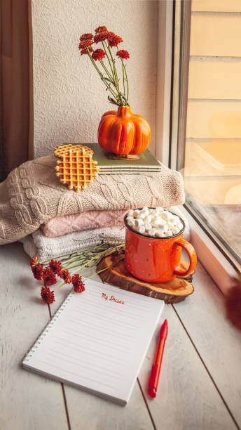Cozy autumn still life on the windowsill with Notepad for notes: warm wool sweaters, pumpkins, maple leaves and a Cup of cocoa with marshmallows and waffles