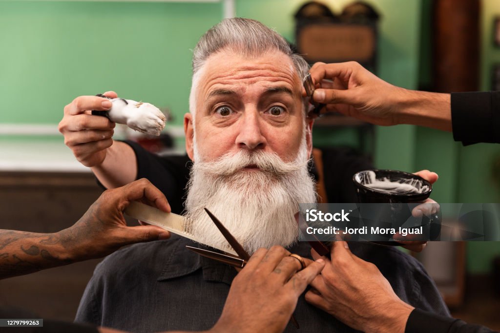 man with white beard in barbershop with barber's hands with cutting and shaving instruments mature man with white beard looking at camera in barber shop with barber hands with cutting and shaving instruments, brush, scissors, comb, razor. hipster style Men Stock Photo
