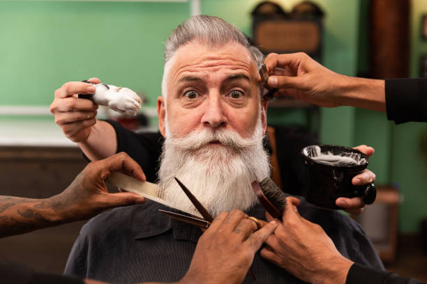 man with white beard in barbershop with barber's hands with cutting and shaving instruments mature man with white beard looking at camera in barber shop with barber hands with cutting and shaving instruments, brush, scissors, comb, razor. hipster style cutting hair stock pictures, royalty-free photos & images