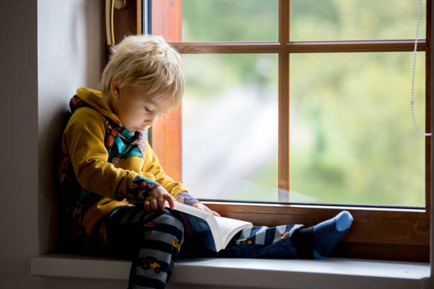istock Cute toddler child, blond boy, sitting on window, reading 1279800486