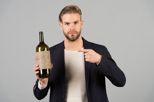 You should try this. Man holds bottle alcohol drink. Social and cultural aspects of drinking. Businessman formal suit confidently welcomes grey background. Man hospitable presenting bottle alcohol.