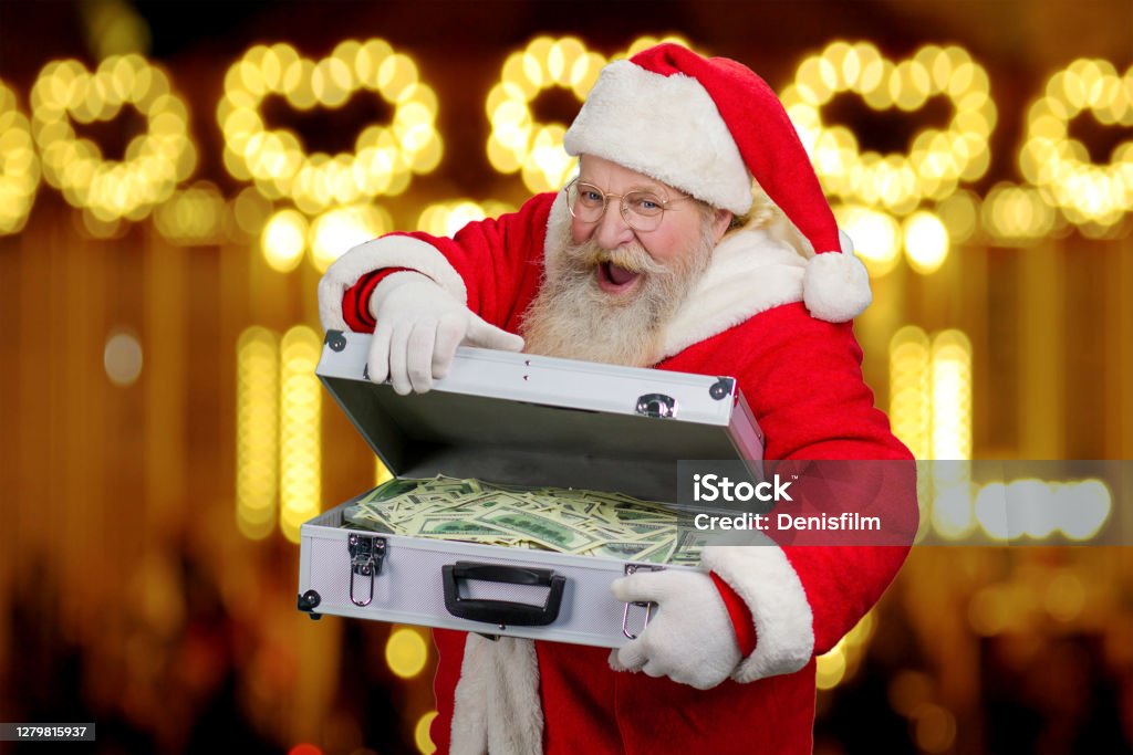 Happy Santa Claus on blurred background. Happy Santa Claus on blurred background. Senior cheerful Santa Claus is opening silver diplomat with dollar bills, New Year lights background. New Year gift. Currency Stock Photo