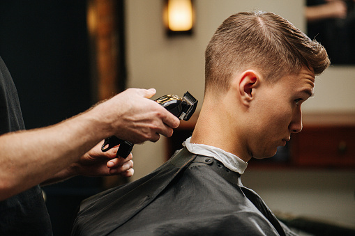 Male hairdresser making a haircut for a young man in a barber shop. He's standing behind client, finishing work with hair clipper. Close up, cropped.