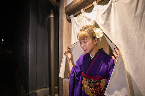 Young woman in ‘Furisode’ kimono walking through ‘Noren’ entrance of traditional Japanese ‘Ryokan’ hotel at night
