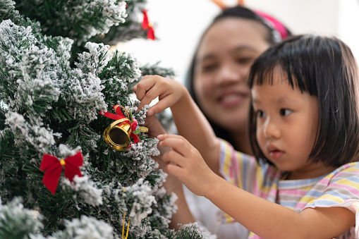 Close up hand of little girl decorating a Christmas tree with mom in background preparing for season greeting of Merry Christmas Happy Holidays. Multigenerational Family engage and happiness concept.