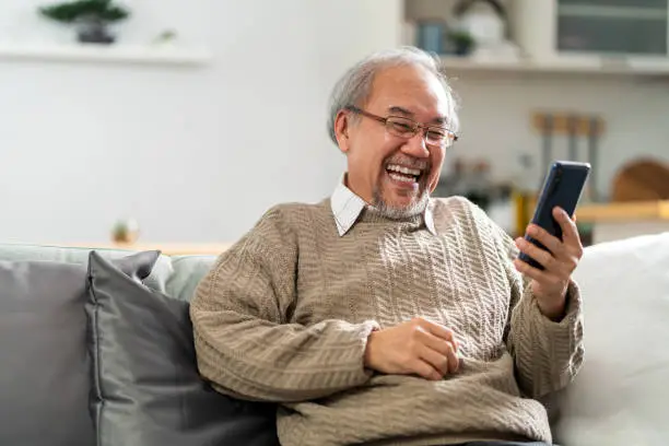 Happy retirement elderly man sitting on sofa at living room use cellphone to make video call to his family with laugh and smile. Communication Technology with senior people at home.