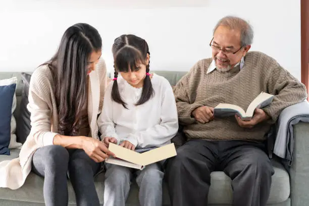 Asian cute girl reading story cartoon book with her mom in living room with grandfather reading fiction book in background. Happiness multigenerational family and domestic lifestyle concept.