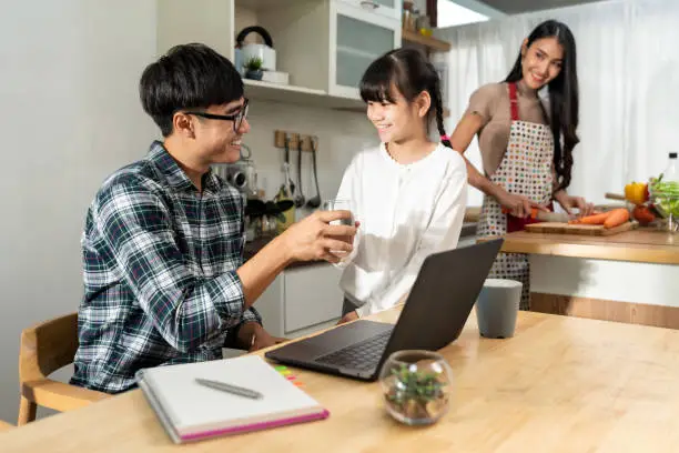 Asian little girl serving water to her dad while working with computer laptop and mom cooking in kitchen. Family Work at home while quarantine from COVID-19. Happy family togetherness concept.
