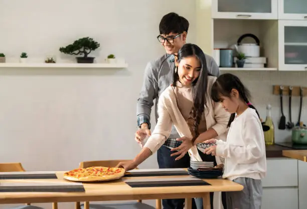 Daughter little girl help mom and dad to setting up dining table before meal. Asian happy family doing domestic life together.