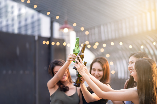 Group of young cute Asian girl gang best friends having party at rooftop restaurant. Girls drinking beer alcohol and toasting bottle feeling happy enjoy, fun with smile face. Night lifestyle concept.