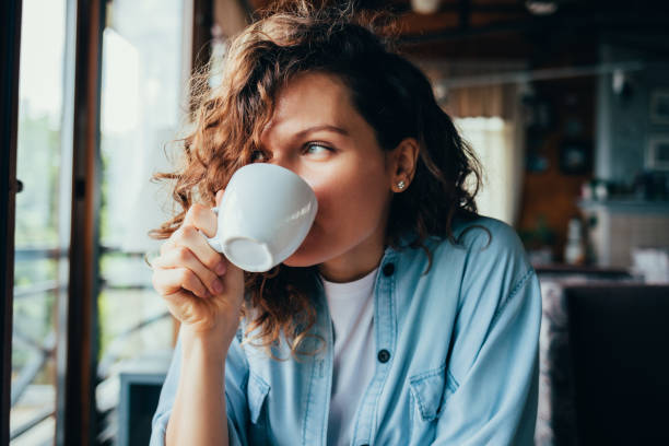 Portrait of happy beautiful young woman Portrait of happy beautiful young woman wearing blue shirt drinking coffee sitting at table in restaurant looking out the window on summer day. coffee drink stock pictures, royalty-free photos & images