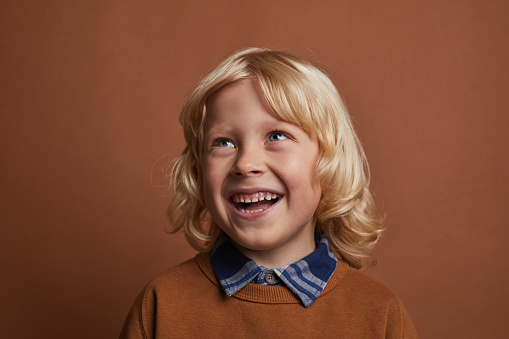 Happy little boy with blond hair wearing shirt and pullover standing against the brown background