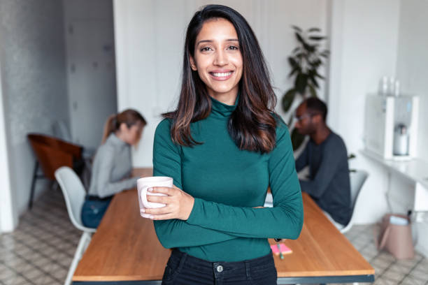 Young indian business woman entrepreneur looking at camera in the office. Shot of young indian business woman entrepreneur looking at camera in the office. In the background, her colleagues working. Best Coffee stock pictures, royalty-free photos & images