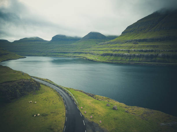 faroe islands kunoy island empty coastal road along the grass covered fjord hills. aerial drone point of view towards the cloud covered mountain peaks and north atlantic ocean horizon. kunoy island, faroe islands, kingdom of denmark, nordic countries, europe