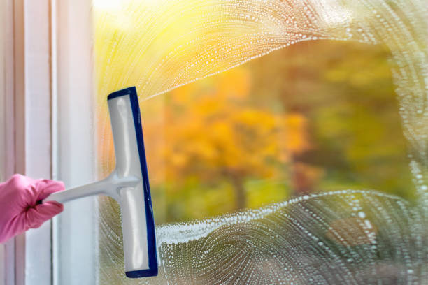 Home routine, cleanliness concept. A woman clean a window pane with a squeegee and soap suds. Hands in pink protective gloves cleaning glass on the windows with a detergent spray bottle. Copy space.