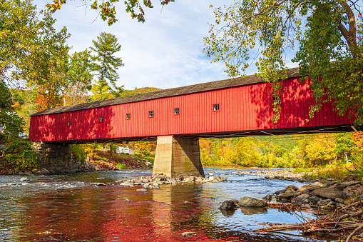 A historic red covered bridge crosses the Housatonic River in West Cornwall, Connecticut on a sunny fall day.