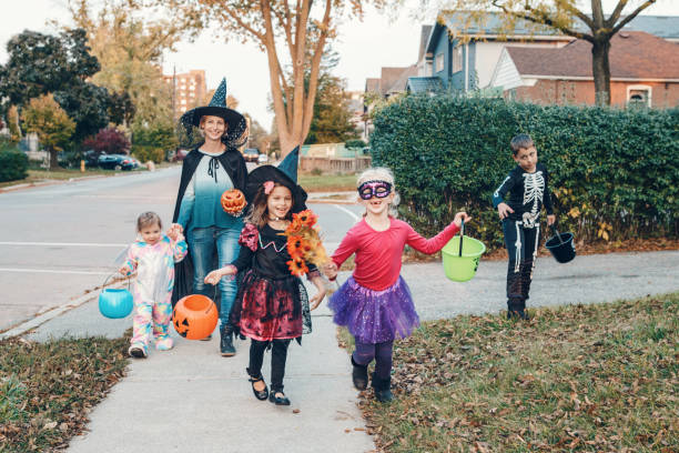 お菓子をくれないといたずらするよ。ハロウィーンの休日にトリックや治療に行く子供を持つ母親。キャンディー、お菓子のために近所の家に行くバスケットとパーティーの衣装で子供を持� - 6 11 months ストックフォトと画像