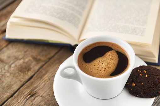 Cup of coffee with book on a wooden table.Reading a book with a cup of espresso coffee on an old vintage wooden table. Cup of coffee against the background of an expanded book.