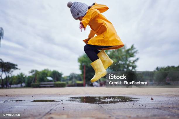 Midair Shot Of A Child Jumping In A Puddle Of Water Wearing Yellow Rubber Boots And A Raincoat In Autumn Stock Photo - Download Image Now