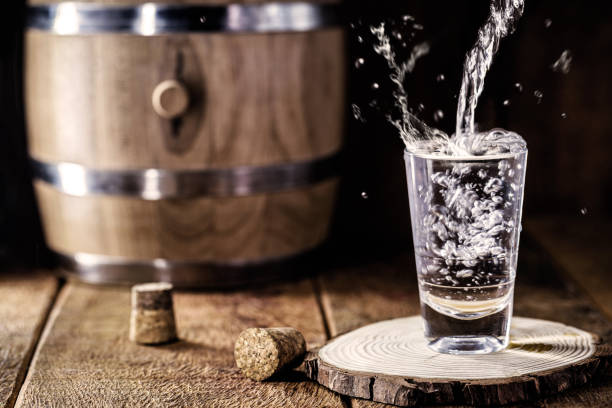 蒸留されたアルコールをショットグラスに注ぎ、飛んで落とし、木製の板の上で提供されます。周りのオーク樽と木製ストッパーとヴィンテージベンチ - whisky alcohol pouring glass ストックフォトと画像