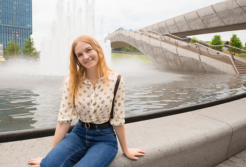 Beautiful young woman with blonde hair posing near fountain at the city background. Girl relax in the street in a sunny day. Travel and active life concept.