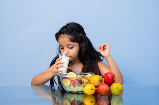 Cute Asian/ Indian little child girl drinking a glass of milk with fruits for breakfast with fun while sitting on dining table. One child person, horizontal, waist up composition with selective focus and copy space.