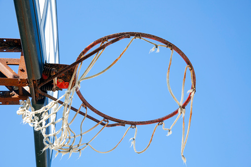 Neglected basketball backboard with rusty hoop and shredded net against blue sky.