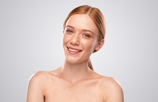 Attractive young female with red hair and freckled skin looking at camera and smiling while representing beauty industry against gray background