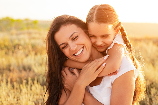 Cheerful little girl embracing smiling woman from back while spending time in field on sunny day together