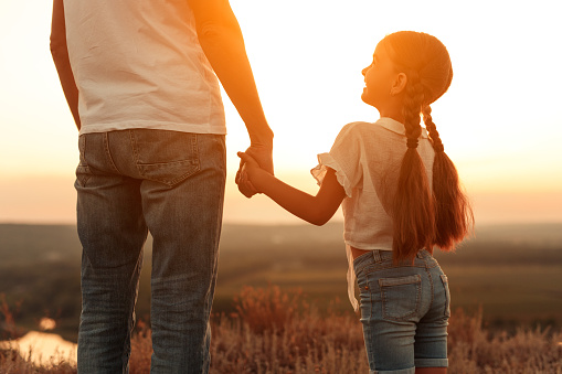 Back view of cheerful girl smiling and holding hand of crop father while admiring sundown in nature together