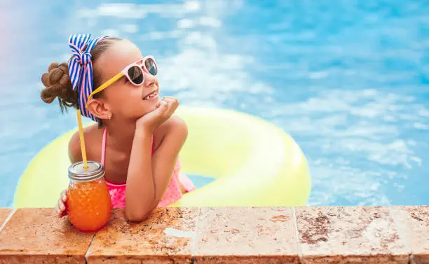 Positive child with jar of fresh fruit drink smiling and looking away while resting in pool near border on summer day on resort