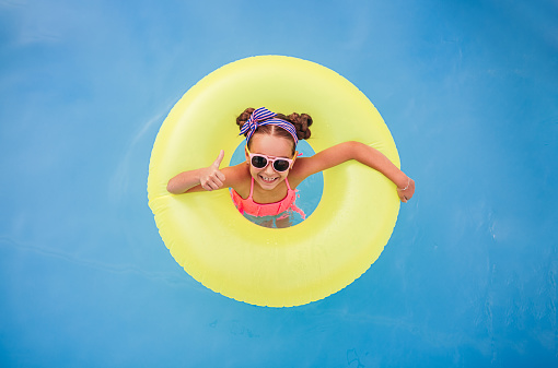 Top view of happy girl in sunglasses swimming in yellow inflatable ring and showing thumb up gesture in pool on resort
