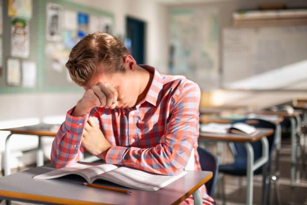 Stressed male student sitting with book at desk. Teenage boy is studying alone in classroom. He is at high school.