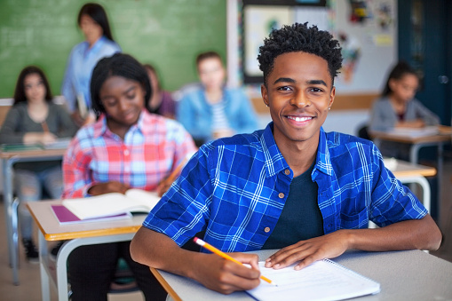 Portrait of smiling male student writing in classroom. Teenage boy is concentrating at high school. He is sitting at desk.