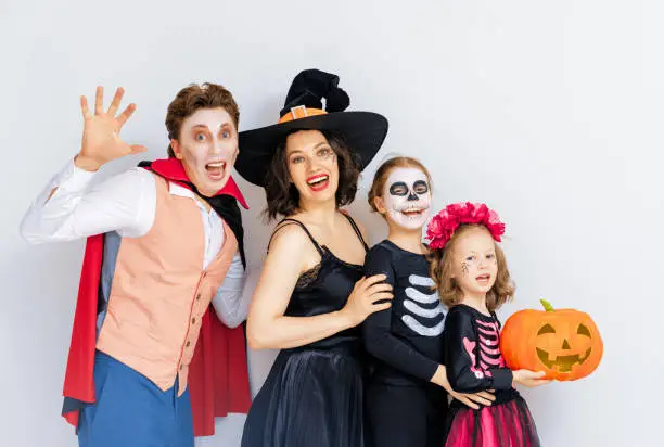 Mother, father and their kids having fun at party. Happy family celebrating Halloween. People wearing carnival costumes and makeup on white wall background.