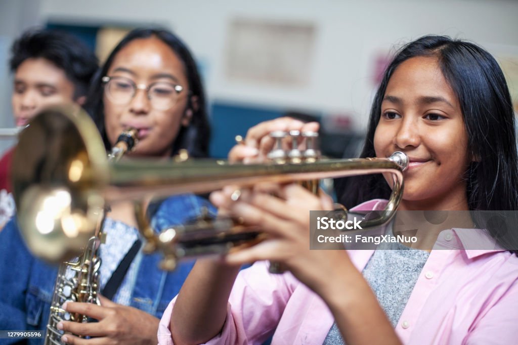 Teenage Asian mixed-ethnicity girl playing trumpet in musical class in school Confident teenage girl playing trumpet in musical class. Female students are learning music at high school. They are practicing woodwind instrument. Playing Stock Photo