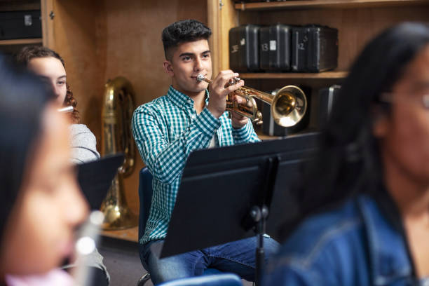 Male Middle Eastern ethnicity student practicing trumpet with friends in school orchestra band Confident male student playing trumpet in musical class. Aspiring teenage boy is learning wind instrument. He is practicing with friends in high school. Music colleges stock pictures, royalty-free photos & images